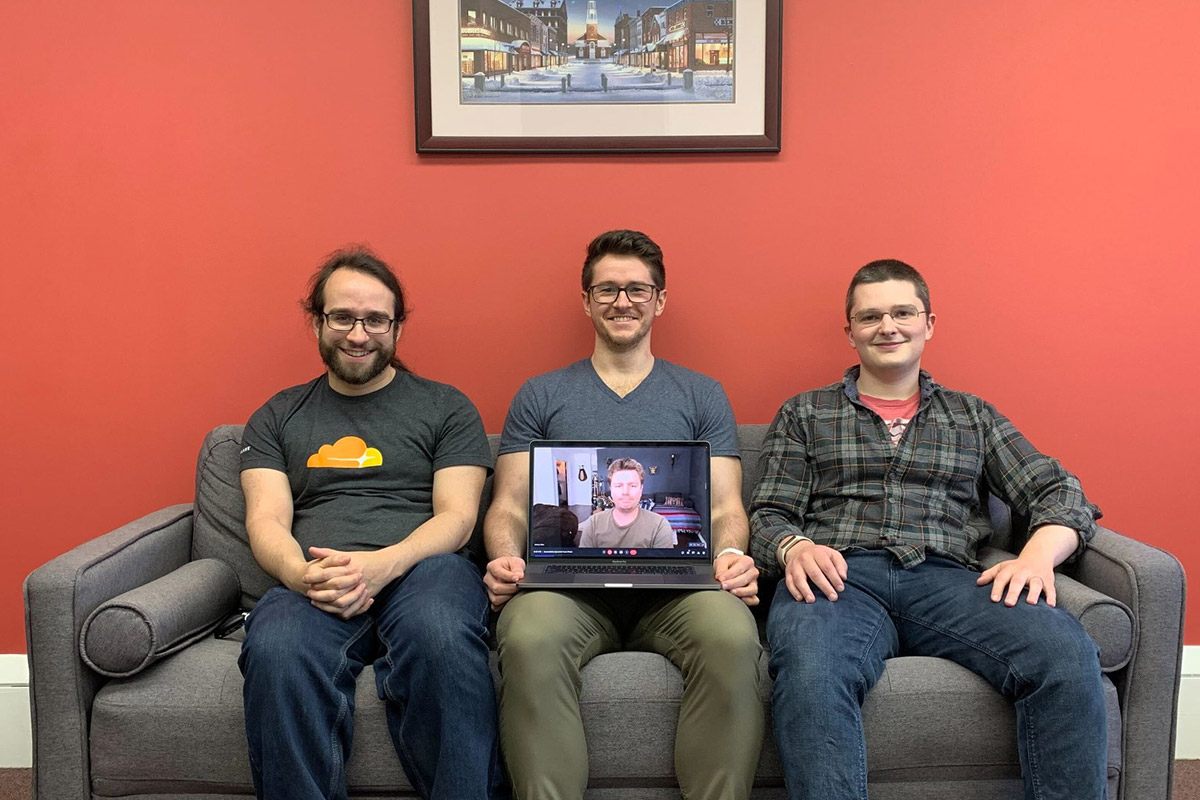 Three of Bytes.co web developers are sitting on a couch and one is holding an open laptop showing a video chat call with one of the remote Bytes.co developeres