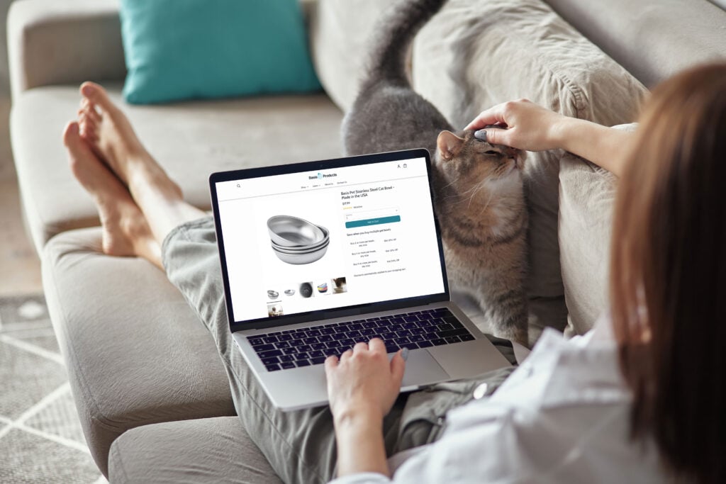 Woman sitting on her couch and petting her cat while browsing for cat bowls on the Basis Products website