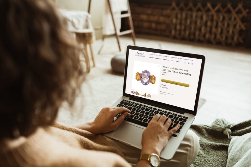 A woman sitting on her couch and using her laptop to shop for a amethyst and citrine ring on the Lippa's Estate & Fine Jewelry website