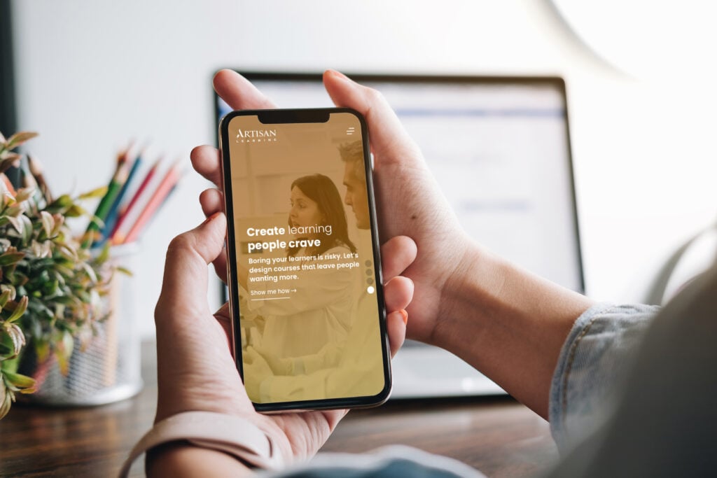 Someone sitting at their desk holding an iPhone with the new Artisan Learning homepage on its screen