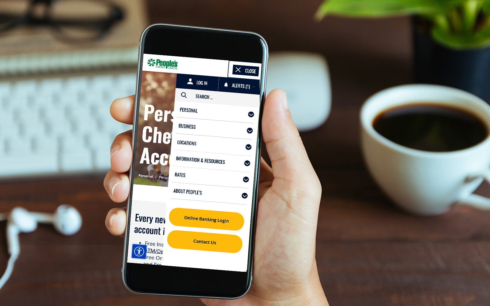 Someone sitting at a desk holding an iPhone with the People's Credit Union website menu open on its screen