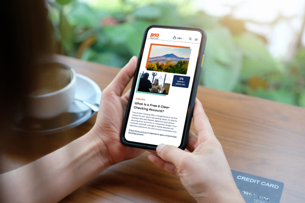 A woman sitting with her coffee and credit card looking at the 802 Credit Union Free & Clear Checking Account page on an iPhone